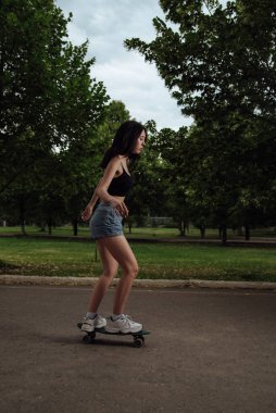 Şehirde kaykay süren genç bir kız. Siyah kolsuz tişörtlü ve mavi şortlu genç bir kadın şehirde kaykayla geziyor. Açık havada kaykay süren genç bir kadın.