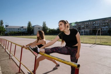 Siyah eşofmanlı iki genç bayan stadyumda antrenman yapıyor. Stadyum arka planda. Kızlar egzersiz yapıyor, geriniyor, yoga yapıyor. Spor ve sağlıklı yaşam tarzı.