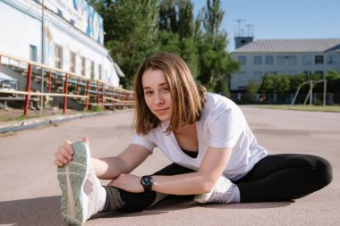 Siyah eşofmanlı genç atletik kadın gün batımında dışarıda antrenman yapıyor. Stadyum arka planda. Kız egzersiz yapıyor, esneme hareketleri yapıyor, yoga yapıyor. Spor ve sağlıklı yaşam tarzı.