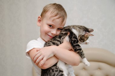 Küçük çocuk kediye sarılıyor. Gülümseyen mutlu küçük çocuk elinde kedi tutuyor. Çocuklar evcil hayvanlarla oynuyor. Çocuklar ve evcil hayvanlar.