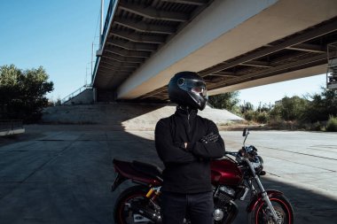 Motosikletli bir erkek motosikletçinin yanında duruyor. Kasklı genç bir adam motosikletin yanında poz veriyor.