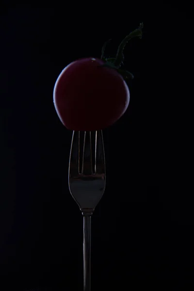 A red cherry tomato with a green stem is put on the tip of a fork against a black background.