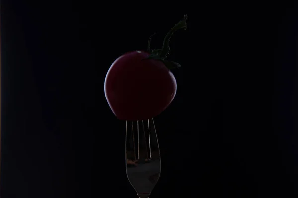 A red cherry tomato is put on the tip of a fork on a black background.