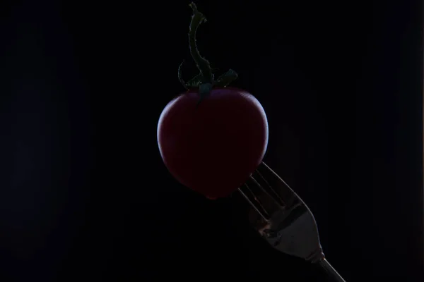 A red cherry tomato is put on the tip of a fork on a black background.