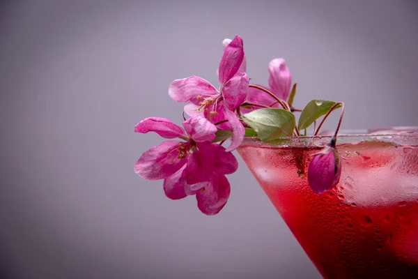 A red alcoholic drink with ice is poured into a martini glass, the glass is decorated with a small branch of a blossoming spring tree with pink flowers.