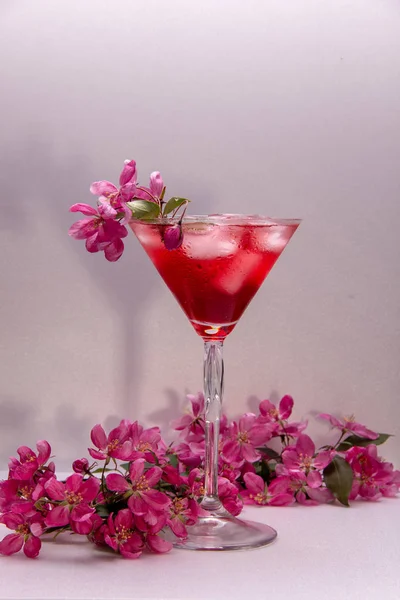 A red sweet drink with ice in a martini glass is decorated with a small branch of a blossoming spring tree with pink flowers.