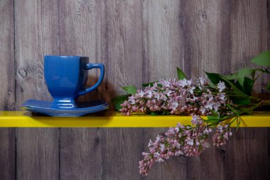 Bir tabak ve mor leylak bir dalı ile mavi bir fincan ahşap bir arka plan üzerinde sarı bir rafta yalan.