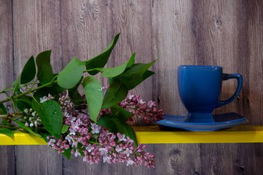 Mavi bir fincan bir tabak üzerinde duruyor, yanında sarı bir rafta çiçekli bahar leylak bir dalı, arkasında ahşap bir arka plan.