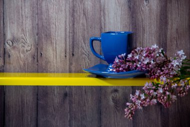 Mavi bir fincan bir tabak üzerinde duruyor, yanında sarı bir rafta çiçekli bahar leylak bir dalı, arkasında ahşap bir arka plan.
