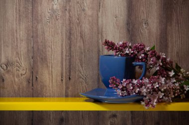 Mavi bir fincan, tabak ve sarı bir rafta çiçek açan bahar leylak standı dalı, arkasında ahşap bir arka plan.