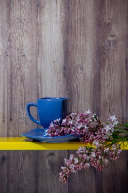 Mavi bir fincan bir tabak üzerinde duruyor, yanında sarı bir rafta çiçekli bahar leylak bir dalı, arkasında ahşap bir arka plan.