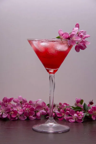 A red alcoholic drink with ice is poured into a martini glass and stands on a wooden table, the glass and table are decorated with branches of a spring flowering tree with pink flowers.