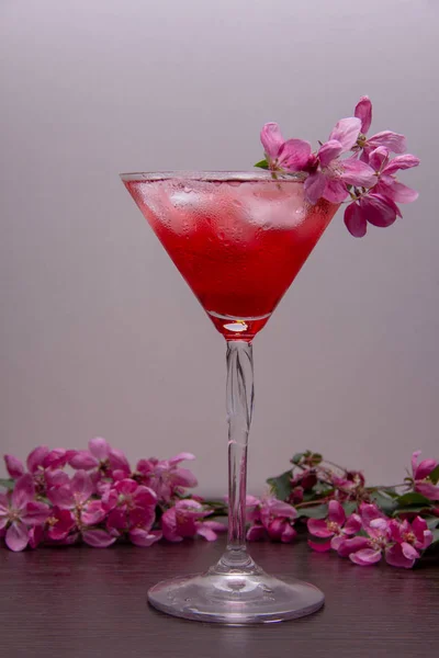 A red alcoholic drink with ice is poured into a martini glass and stands on a wooden table, the glass and table are decorated with branches of a spring flowering tree with pink flowers.