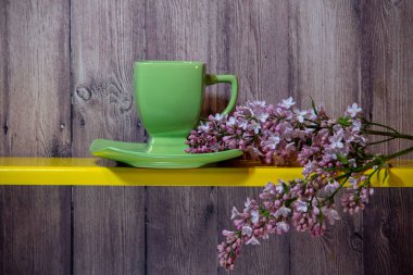 Yeşil bir fincan, tabak ve sarı bir rafta çiçek açan bahar leylak standı dalı, arkasında ahşap bir arka plan.
