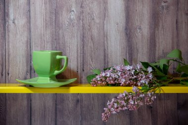 Yeşil bir fincan, tabak ve sarı bir rafta çiçek açan bahar leylak standı dalı, arkasında ahşap bir arka plan.