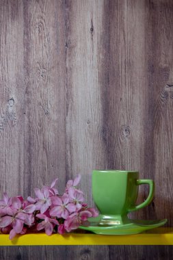 Yeşil fincan, tabak, sarı rafta pembe çiçekler ile çiçekli dal, ahşap bir arka plan arkasında.