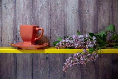 Kırmızı bardak ve tabak sarı bir rafta, bir leylak çiçekli dalı yanında, ahşap bir arka plan arkasında.