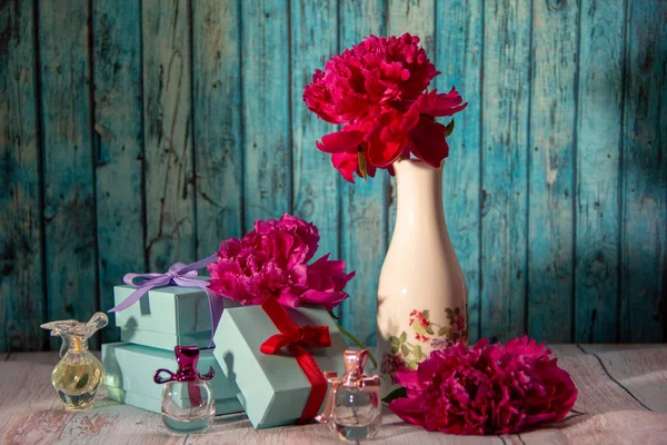 Three small gift boxes decorated with bows, three small bottles of women\'s perfume, one red peony in a white vase with painted flowers, two peony flowers on a blue wooden background.