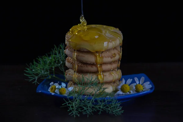 Five round biscuits stand on top of each other, blooming daisies and green grass lie next to the biscuits on a blue saucer, honey flows from the top of the biscuits.