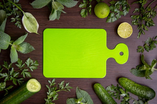 Green cutting board lies in the center of the table, around are ripe cucumbers, fragrant limes, parsley branches, fresh mint branches.