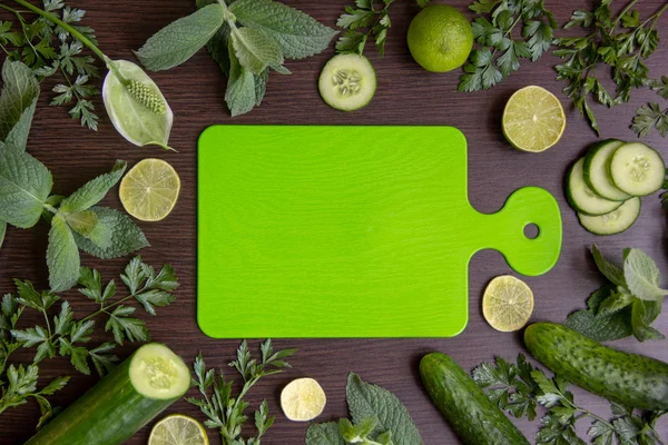 The green cutting board lies in the center of the table, around are ripe cucumbers, fragrant limes, parsley branches, fresh mint branches, lime slices and cucumber.