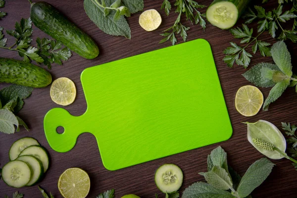 Green cutting board lies in the center of the table, around are ripe cucumbers, fragrant limes, parsley branches, fresh mint branches.