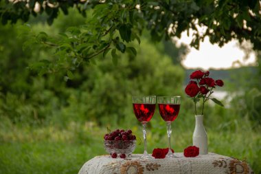 Kırmızı bir içecek ile iki bardak, olgun kiraz ile kristal vazo, kırmızı gülile bir vazo doğada yeşil bir bahçede güzel bir masa örtüsü ile dekore edilmiş bir masa üzerinde durmak.