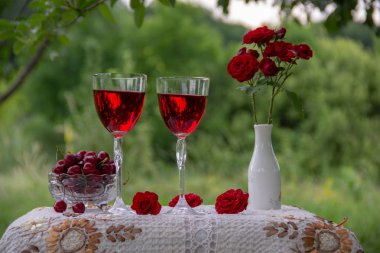 Kırmızı içecek ile iki bardak, olgun kiraz ile kristal vazo, kırmızı gülile bir vazo, yeşil bir bahçede güzel bir masa örtüsü ile dekore edilmiş bir masa üzerinde hediye standı ile bir kutu.