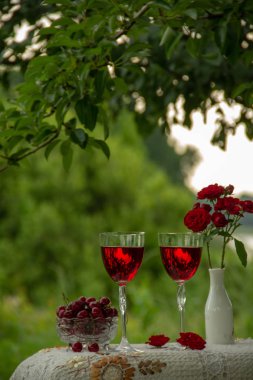 Kırmızı bir içecek ile iki bardak, olgun kiraz ile kristal vazo, kırmızı gülile bir vazo doğada yeşil bir bahçede güzel bir masa örtüsü ile dekore edilmiş bir masa üzerinde durmak.