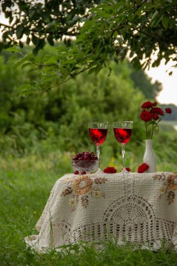 Kırmızı bir içecek ile iki bardak, olgun kiraz ile kristal vazo, kırmızı gülile bir vazo doğada yeşil bir bahçede güzel bir masa örtüsü ile dekore edilmiş bir masa üzerinde durmak.