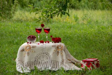 Kırmızı bir içecek ile iki bardak, kokulu gül bir buket, güzel bir masa örtüsü ile dekore edilmiş bir masa üzerinde kristal vazo standında olgun kiraz, yeşil bir yaz bahçesinde bir hediye ile bir kutu yanında.