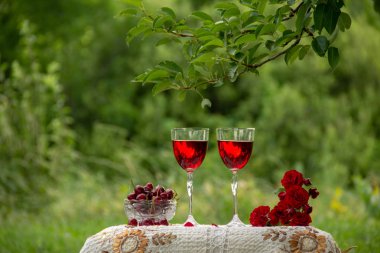 Kırmızı bir içecek ile iki bardak, kokulu gül bir buket, yeşil bir yaz bahçesinde güzel bir masa örtüsü ile dekore edilmiş bir masa üzerinde kristal vazo standında olgun kiraz.