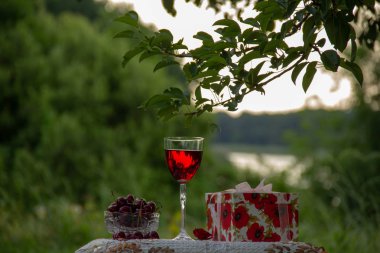 Bir kadeh kırmızı şarap, kristal vazoda olgun tatlı kiraz, ağaçların arasında yeşil bir bahçede güzel bir masa örtüsü yle süslenmiş bir masanın üzerinde hediye standı olan bir kutu.