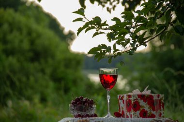 Kırmızı bir içecek ile bir bardak, kristal vazo olgun bir kiraz, doğada yeşil bir yaz bahçesinde güzel bir masa örtüsü ile dekore edilmiş bir masada bir hediye standı ile parlak bir kutu.