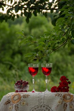 Kırmızı bir içecek ile iki bardak, kokulu gül bir buket, yeşil bir yaz bahçesinde güzel bir masa örtüsü ile dekore edilmiş bir masa üzerinde kristal vazo standında olgun kiraz.