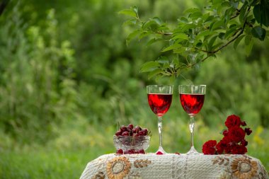 Kırmızı bir içecek ile iki bardak, kokulu gül bir buket, yeşil bir yaz bahçesinde güzel bir masa örtüsü ile dekore edilmiş bir masa üzerinde kristal vazo standında olgun kiraz.
