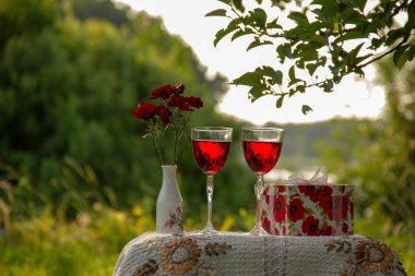 Kırmızı içecekli iki bardak, vazoda güller, ağaçların arasında yeşil bir bahçede güzel bir masa örtüsüyle süslenmiş bir masanın üzerinde hediye standı olan bir kutu..