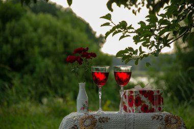 Kırmızı içecekli iki bardak, vazoda güller, ağaçların arasında yeşil bir bahçede güzel bir masa örtüsüyle süslenmiş bir masanın üzerinde hediye standı olan bir kutu..