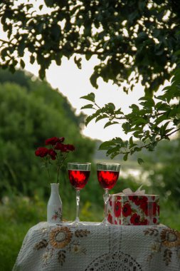 Kırmızı bir içecek ile iki bardak, bir vazo kokulu gül bir buket, ağaçların arasında yeşil bir yaz bahçesinde güzel bir masa örtüsü ile dekore edilmiş bir masa üzerinde bir hediye standı ile bir kutu.