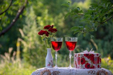 Kırmızı bir içecek ile iki bardak, bir vazo kokulu gül bir buket, ağaçların arasında yeşil bir yaz bahçesinde güzel bir masa örtüsü ile dekore edilmiş bir masa üzerinde bir hediye standı ile bir kutu.