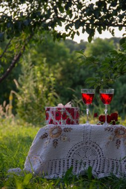 Kırmızı bir içecek ile iki bardak, kokulu gül bir buket, doğada yeşil bir yaz bahçesinde güzel bir masa örtüsü ile dekore edilmiş bir masa üzerinde bir hediye standı ile bir kutu.