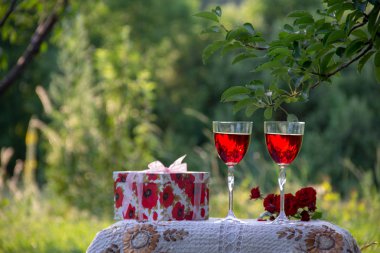 Kırmızı bir içecek ile iki bardak, kokulu gül bir buket, doğada yeşil bir yaz bahçesinde güzel bir masa örtüsü ile dekore edilmiş bir masa üzerinde bir hediye standı ile bir kutu.