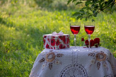 Kırmızı bir içecek ile iki bardak, kokulu gül bir buket, doğada yeşil bir yaz bahçesinde güzel bir masa örtüsü ile dekore edilmiş bir masa üzerinde bir hediye standı ile bir kutu.