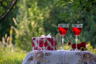 Kırmızı bir içecek ile iki bardak, kokulu gül bir buket, doğada yeşil bir yaz bahçesinde güzel bir masa örtüsü ile dekore edilmiş bir masa üzerinde bir hediye standı ile bir kutu.