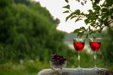 Doğada yeşil bir yaz bahçesinde güzel bir masa örtüsü ile dekore edilmiş bir masa üzerinde kristal vazo standında kırmızı bir içecek ile iki bardak, olgun kiraz.