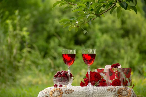 Two glasses with a red drink, a crystal vase with ripe cherries, a bouquet of red roses, a gift box stand on a table decorated with a beautiful tablecloth in a green garden in nature.