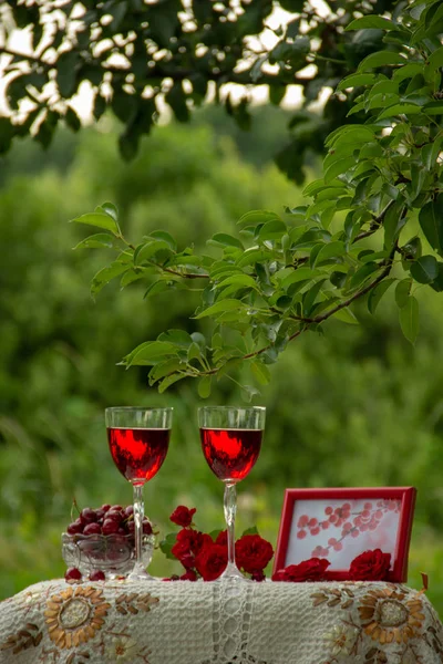 Two glasses with a red drink, a crystal vase with ripe cherries, a bouquet of red roses, a photo frame with a picture, stand on a table decorated with a beautiful tablecloth in a green garden in nature.