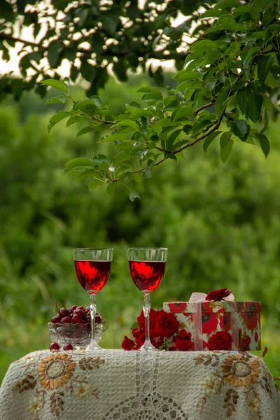 Two glasses with a red drink, a crystal vase with ripe cherries, a bouquet of red roses, a gift box stand on a table decorated with a beautiful tablecloth in a green garden in nature.