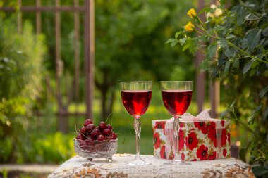 Kırmızı bir içecek ile iki bardak, kristal vazo içinde olgun kiraz, çiçek açan gül ler yakın sokakta güzel bir masa örtüsü ile dekore edilmiş bir masada bir hediye standı ile parlak bir kutu.