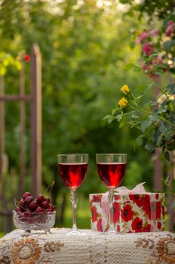 Kırmızı bir içecek ile iki bardak, kristal vazo içinde olgun kiraz, çiçek açan gül ler yakın sokakta güzel bir masa örtüsü ile dekore edilmiş bir masada bir hediye standı ile parlak bir kutu.